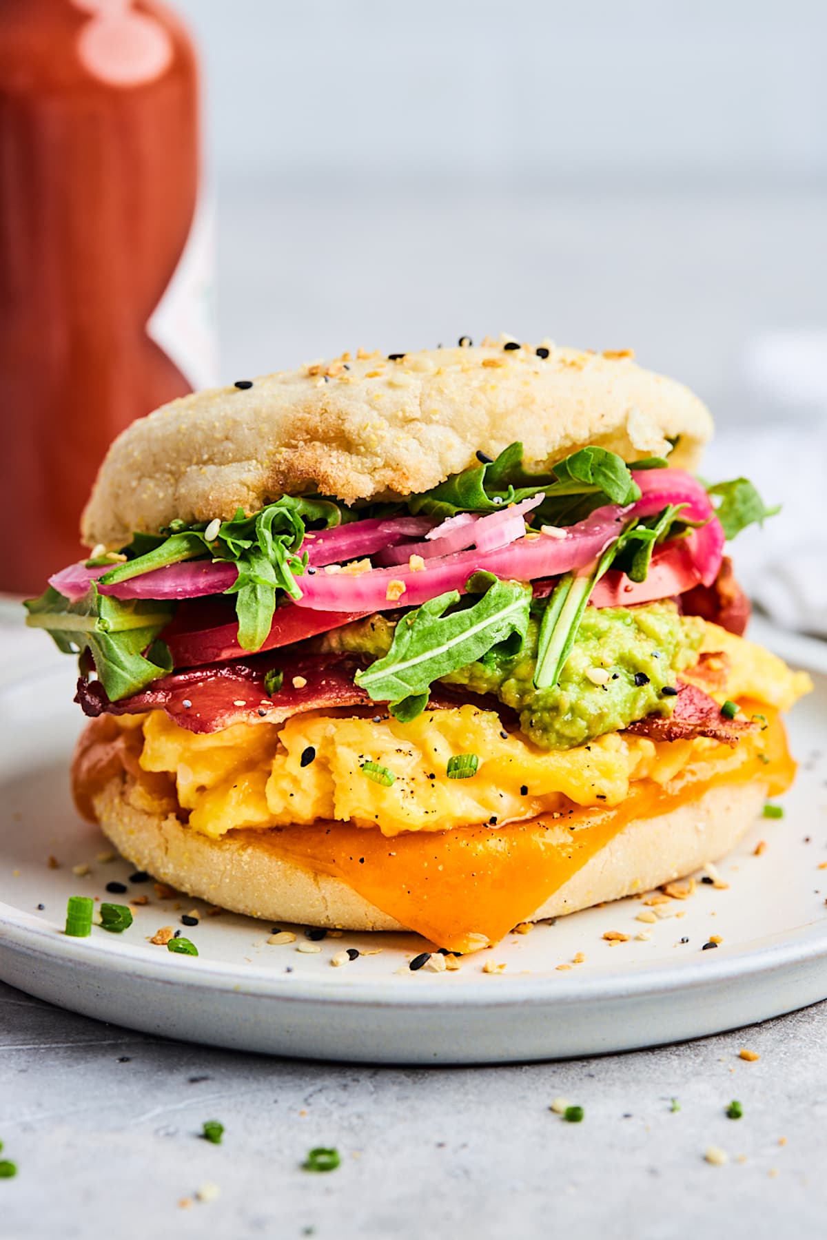 English muffin breakfast sandwich on plate with cheese, scrambled eggs, bacon, avocado, arugula, and pickled red onion. 