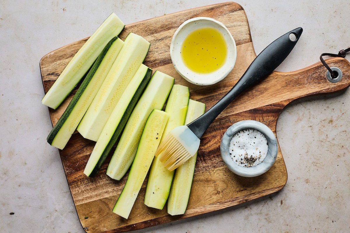 zucchini cut into spears on wood cutting board being brushed with olive oil. 