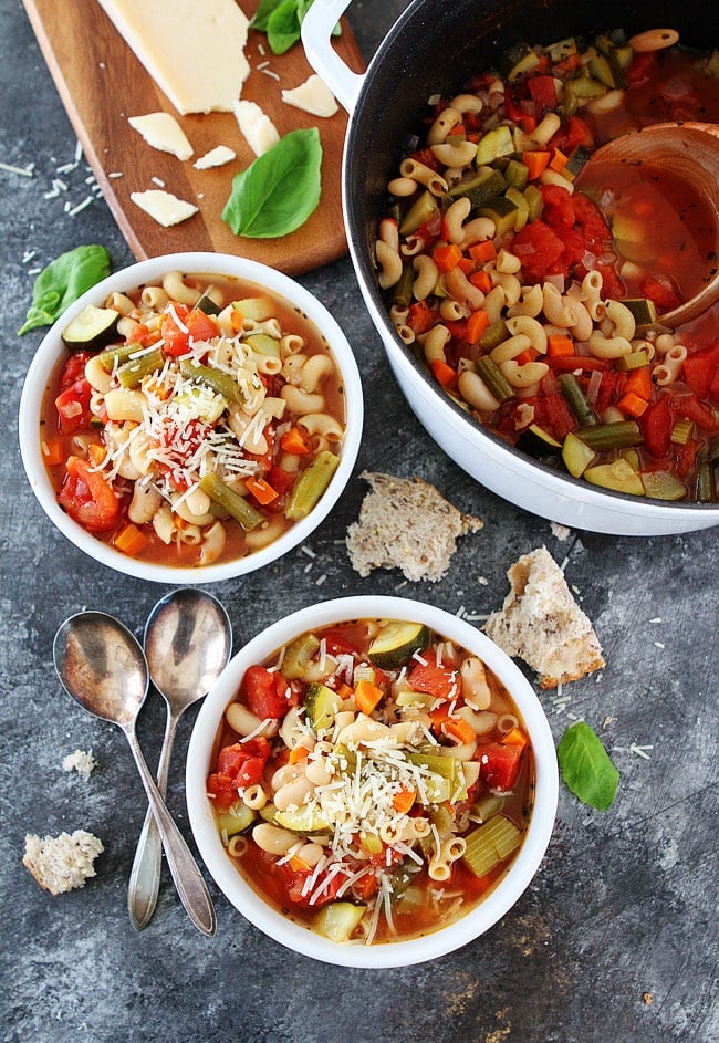 Minestrone soup in two white bowls and large white pot with crusty bread and spoons. 