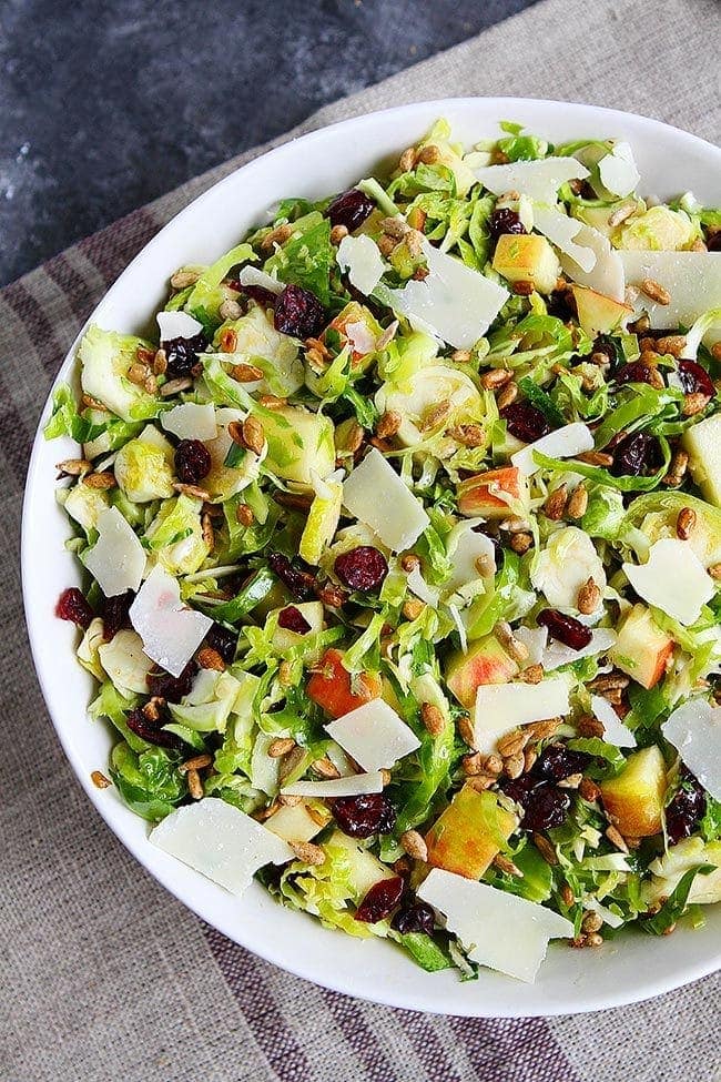 A bowl of shaved Brussels sprouts salad, with large chunks of apple and dried cranberries, topped with shaved parmesan.