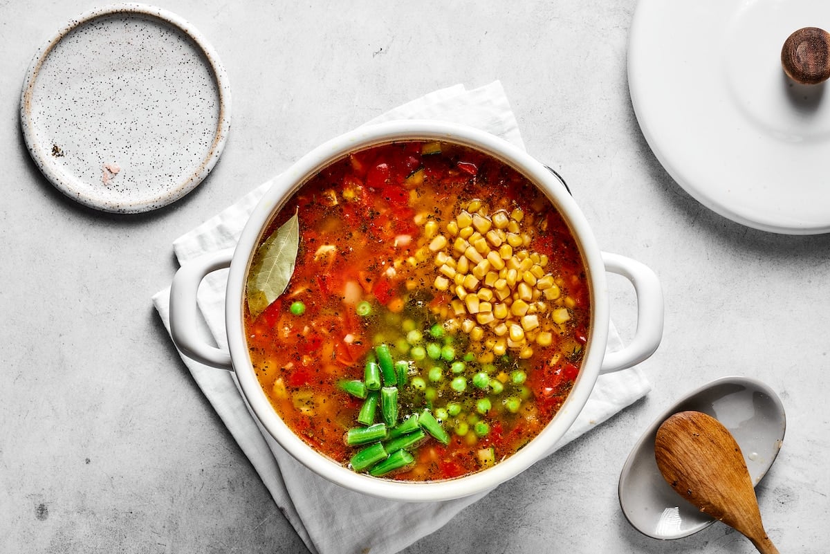 Frozen veggies are added to vegetable soup in a saucepan.