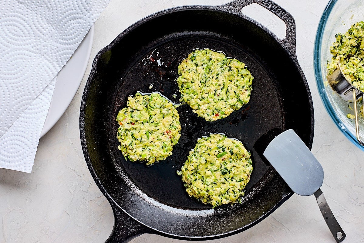 zucchini fritters frying in cast iron skillet.